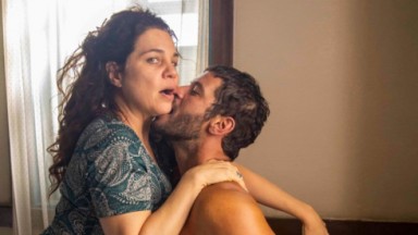 Maria Bruaca (Isabel Teixeira) recebendo um beijo de Levi (Leandro Lima) no pescoço, em cima da cama 