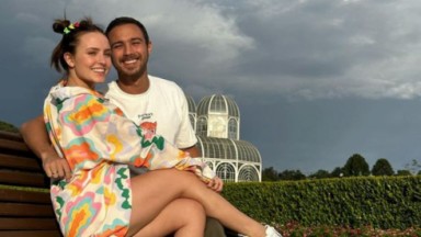 Larissa Manoela e André Luiz Frambach sorrindo, abraçados, sentados em um banco 