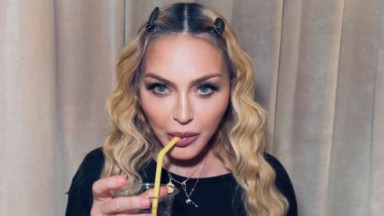 Madonna bebendo algo em copo com canudo amarelo, olhando para a câmera, de camiseta preta 