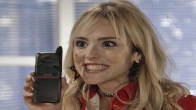 Isabelle Drummond, caracterizada como Manuzita de "Verão 90" sorri segurando um celular flip dos anos 90. 