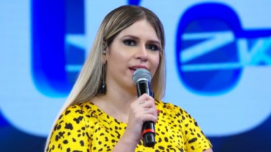 Marília Mendonça em entrevista ao Domingão com Huck. Na foto, ela segura um microfone e usa uma blusa amarela com manchas pretas 