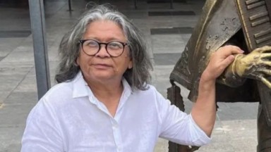 Marlene Mattos de camisa social branca e óculos, posando ao lado de estátua de bronze, sem sorrir 