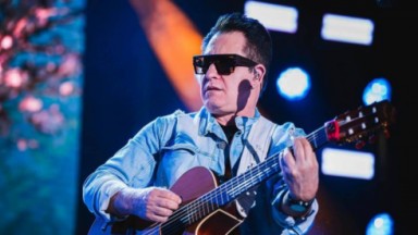 Marrone de roupa jeans, tocando violão, de óculos escuros em palco 