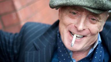 Brian Travers sorridente, com um cigarro na boca 