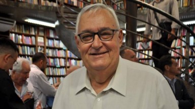 José Nello Marques de camisa social branca, sorrindo em livraria com pessoas atrás, de óculos 