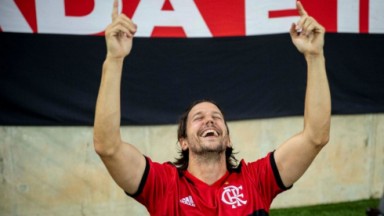 Neném (Vladimir Brichta) vestindo camisa do Flamengo, sorrindo e apontando para o alto com as duas mãos 