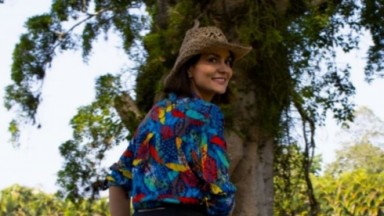 Paula Barbosa de chapéu e camisa colorida sorrindo para foto 
