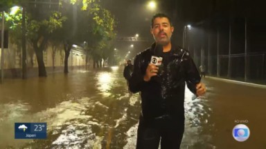 Repórter Chico Regueira molhado, segurando microfone, debaixo de chuva, no meio de uma rua alagada 