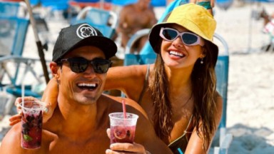 Ricardo Maia e Bárbara Heck posando para foto na praia, segurando copos de drinks, usando óculos escuros e chapéus 