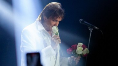 Roberto Carlos de roupa branca, com rosas na mão, perto de microfone, com cabeça abaixada em show 