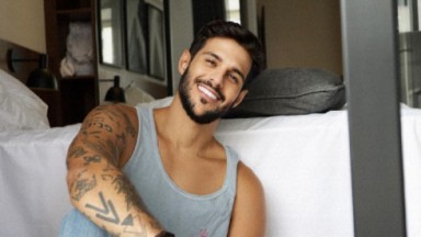 Rodrigo Mussi, de camiseta regata, sorridente, sentado na frente de uma cama 