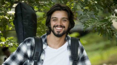 Rodrigo Simas de camisa xadrez e mochila nas costas sorrindo 
