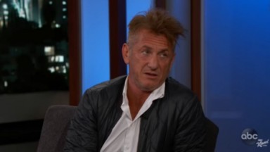 Sean Penn de camisa branca e jaqueta preta em cenário de talk show, com expressão confusa 
