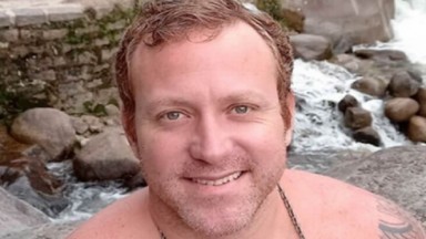 Sérgio Hondjakoff sorridente com uma cachoeira e pedras ao fundo 