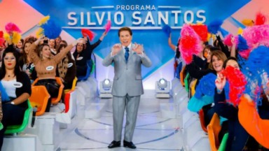 Silvio Santos de terno cinza, camisa branca e gravata azul, sorrindo, com as mãos levantadas 