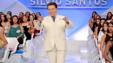 Silvio Santos no palco de seu programa, de roupa bege, sorrindo e apontando para frente 