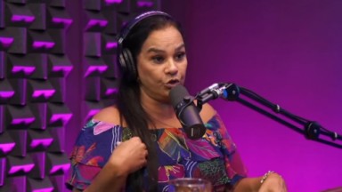 Solange Couto na participação do podcast Papagaio Falante; atriz está sentada, usa blusa estampada, fone e fala em microfone de mesa 