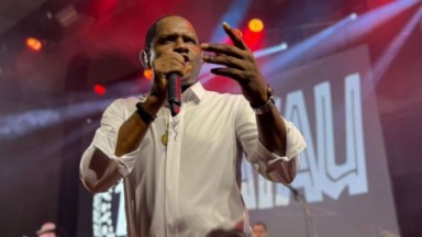 Tatau cantando com microfone, em palco, com camisa social branca 