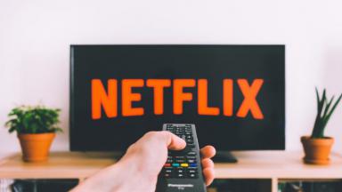 Mão apertando controle remoto da TV, e que aparece o logo da Netflix 