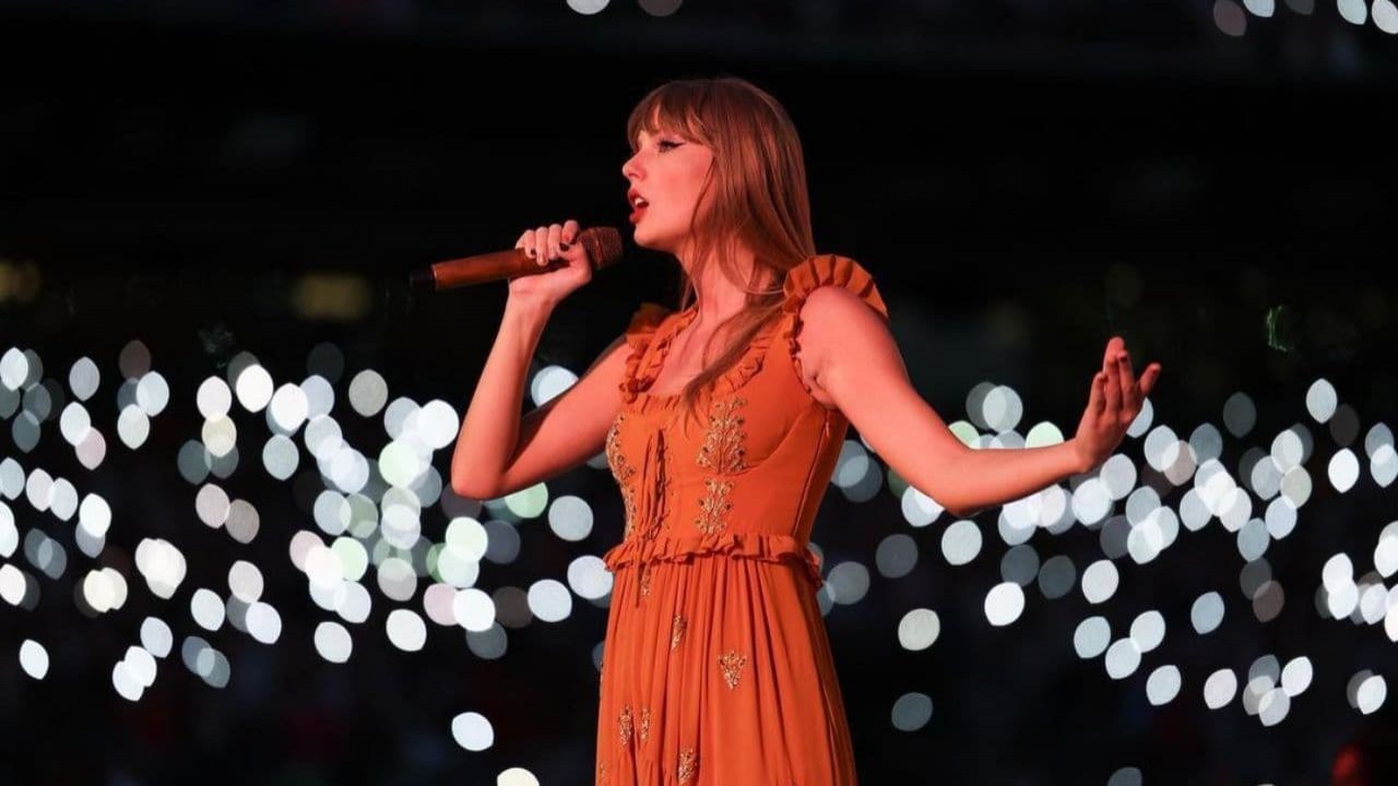 Taylor Swift cantando, com cabelo solto e vestido coral