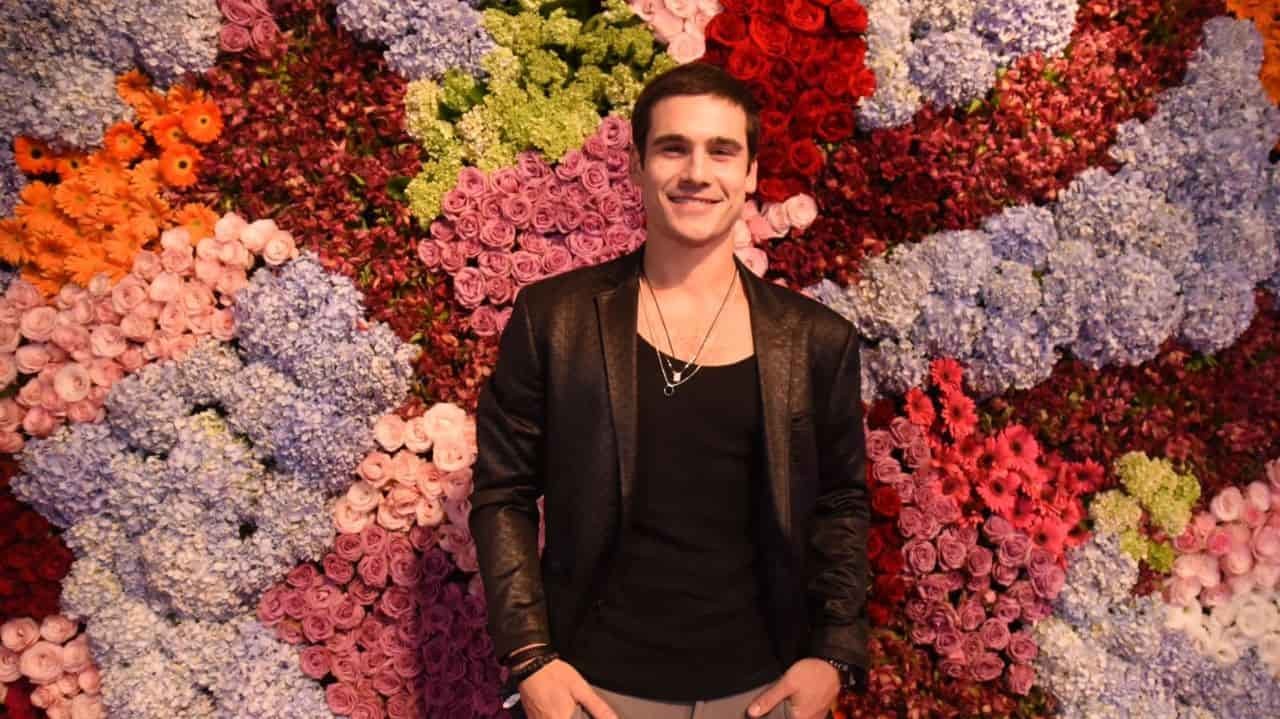 Nicolas Prattes posando e sorrindo próximo a parede de flores coloridas