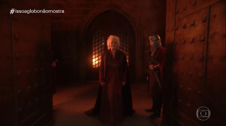 Ana Maria Braga vestindo roupas medievais num cenário que lembra um castelo, ao lado, um guarda.