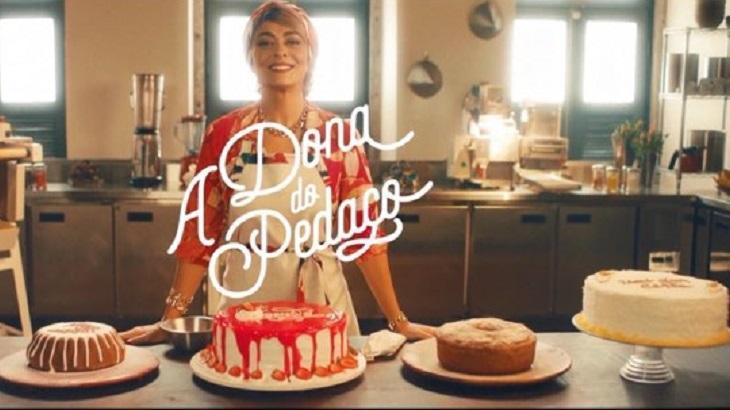 Juliana Paes caracterizada com boleira, numa cozinha, com vários bolos na sua frente. E o logo de "A Dona do Pedaço" na tela