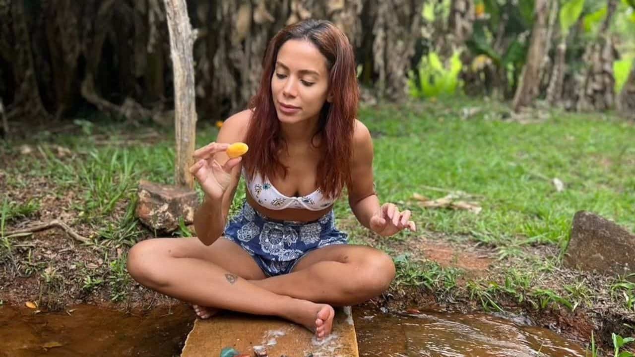 Anitta sentada perto de mato e riacho, com fruta na mão e cabelo solto