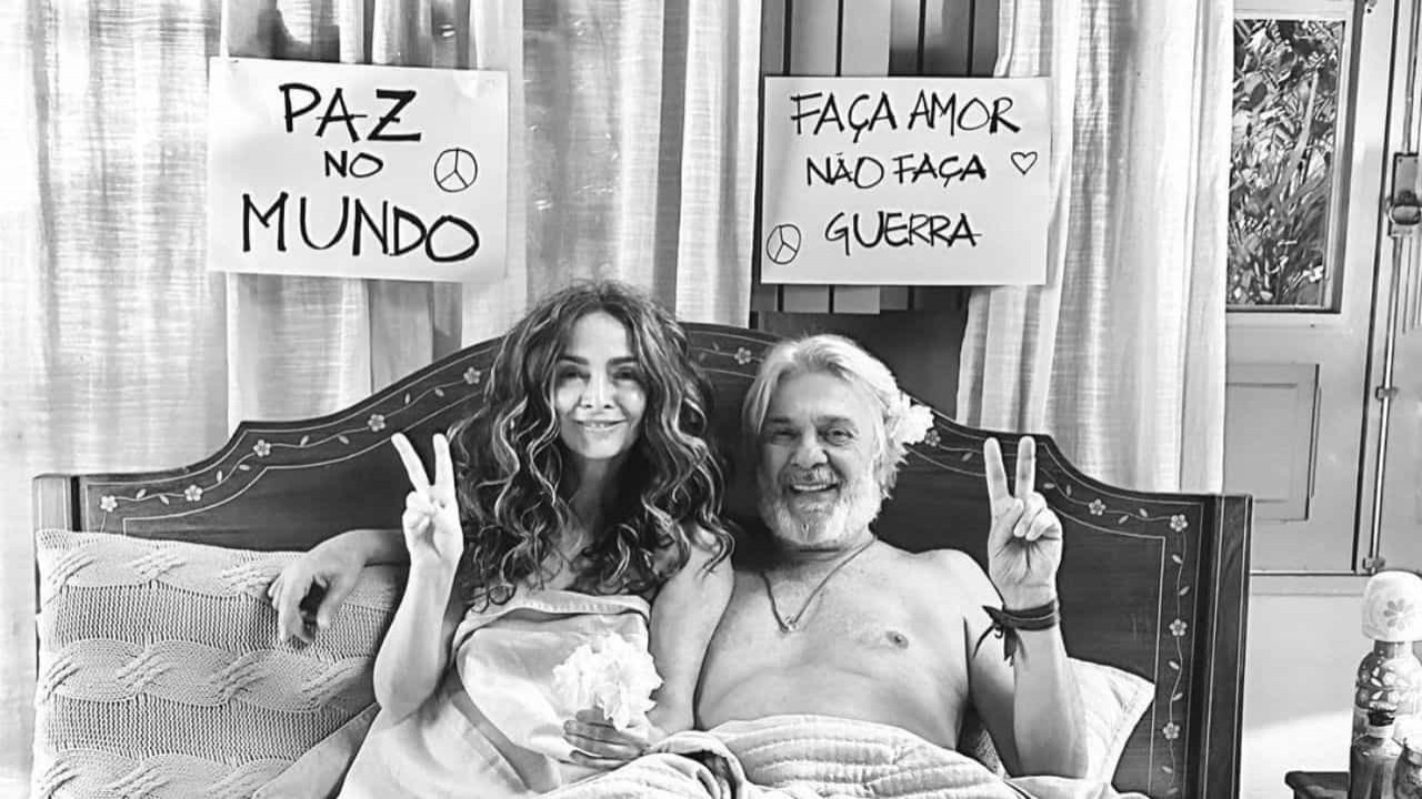 Claudia Ohana e Zé Carlos Machado deitados na cama, cobertos por lençóis, sorrindo e fazendo sinal de paz e amor