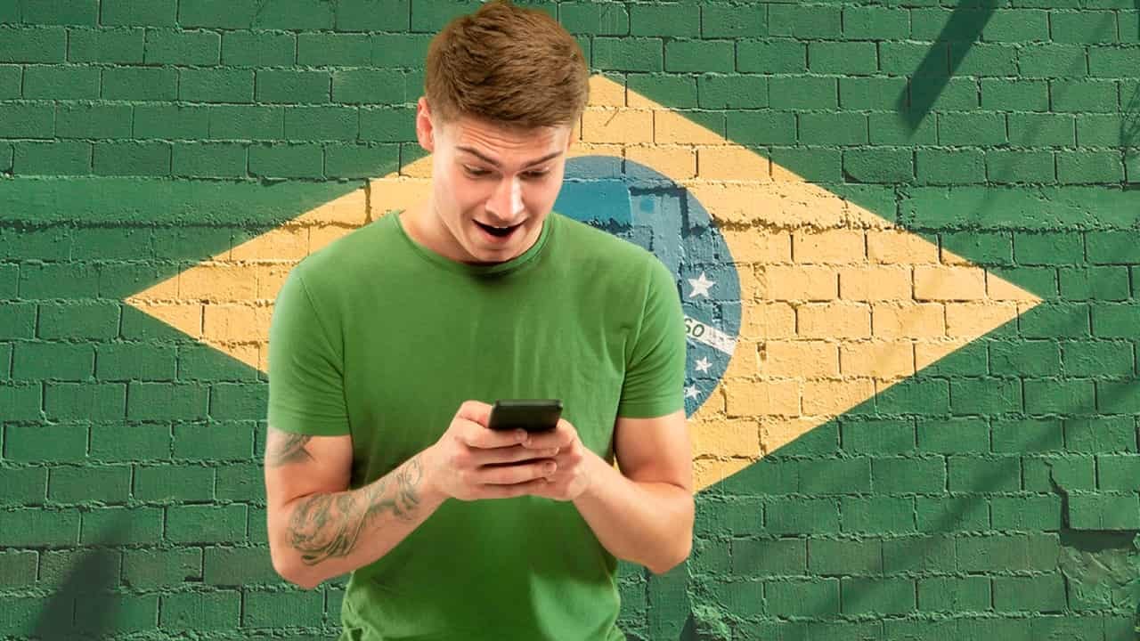 Rapaz com o celular na mão e bandeira do Brasil ao fundo