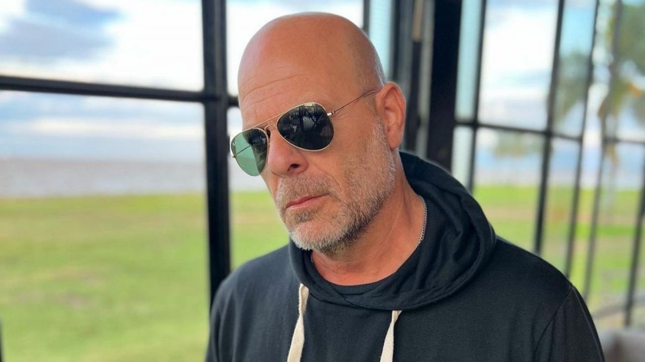 Bruce Willis posando para foto vestindo moletom preto e usando óculos escuros