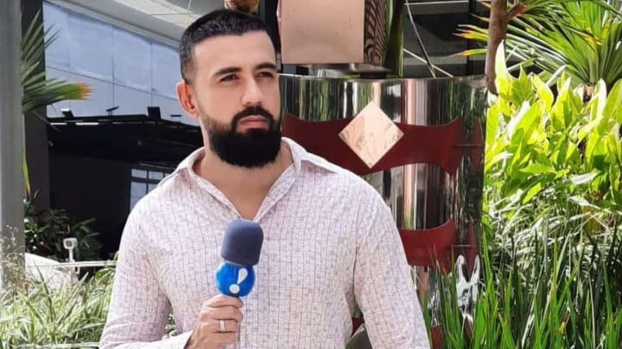 Bruno Tálamo de camisa social clara, segurando microfone da RedeTV! e sem sorrir