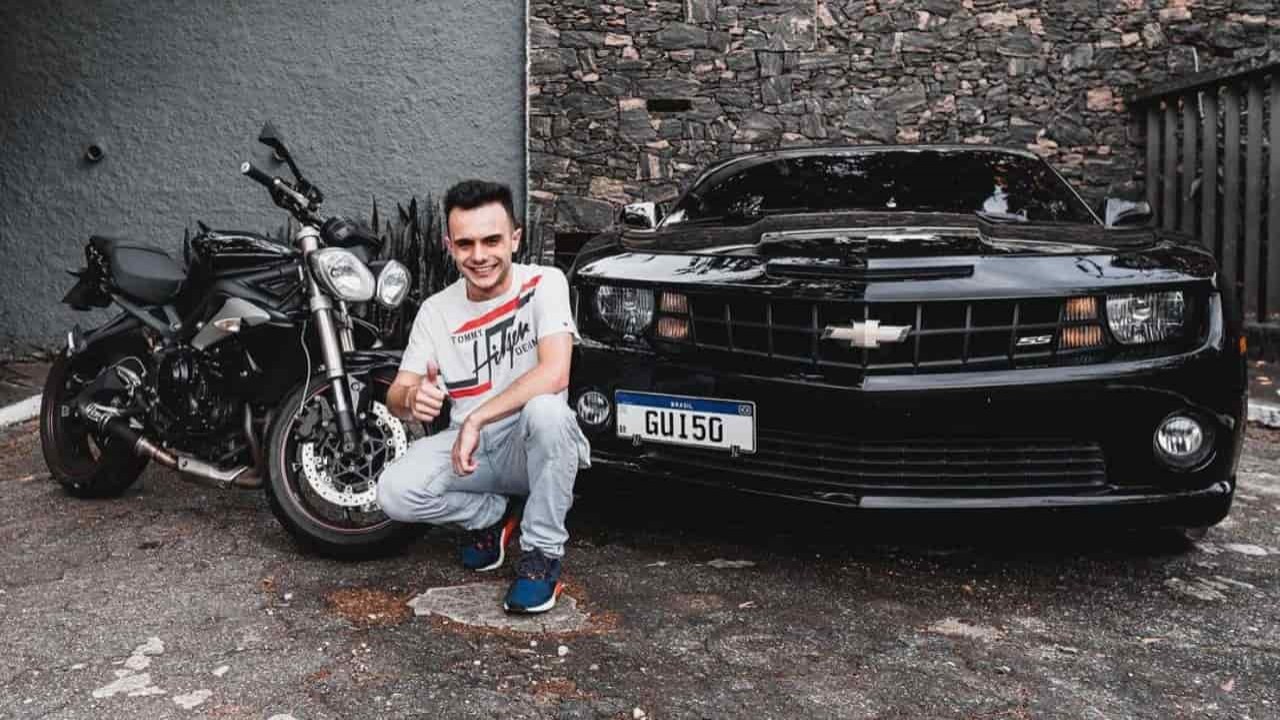 Guillermo Hundadze agachado, sorrindo, perto de carro e moto