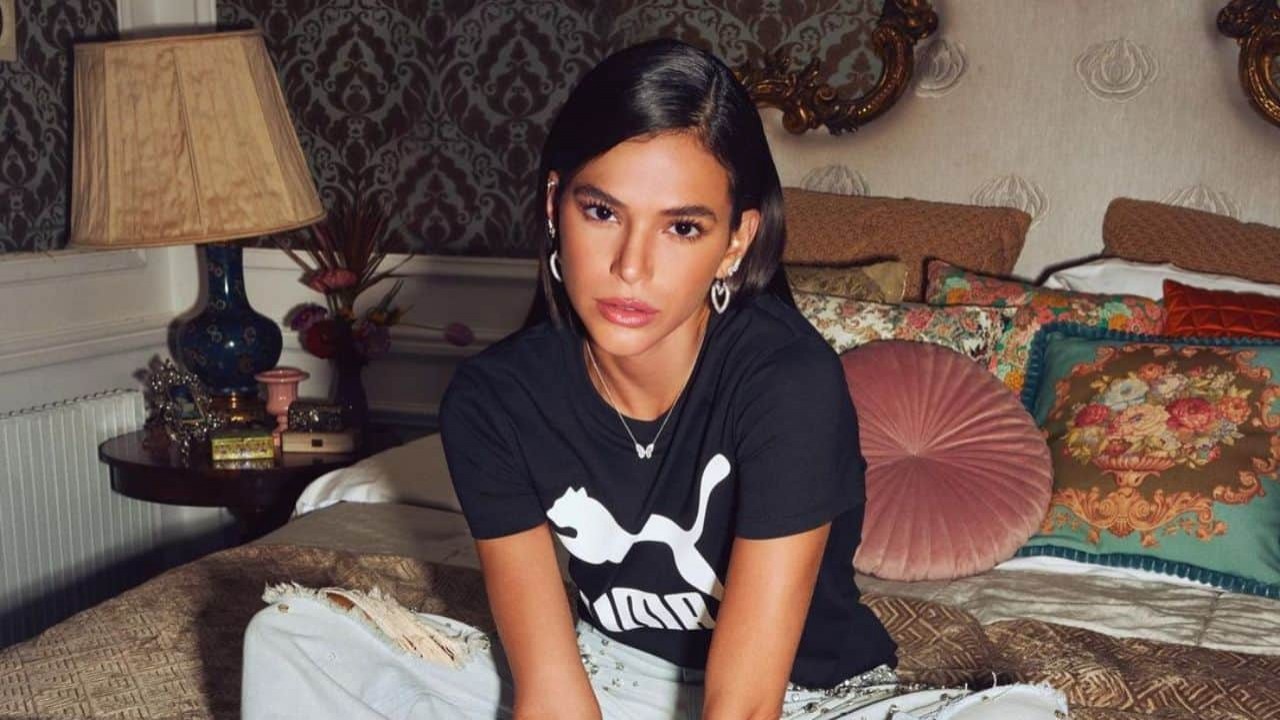 Bruna Marquezine sem sorrir, de cabelos soltos, sentada em cama com blusa preta e calça jeans