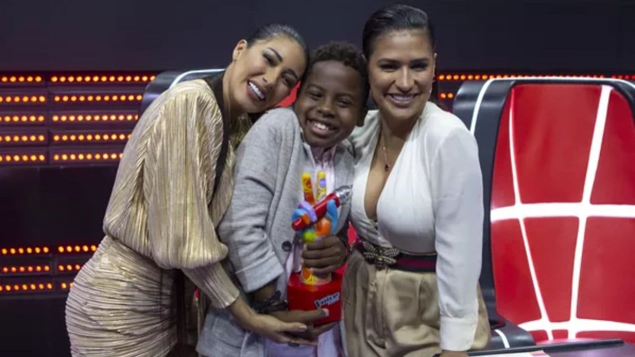 Jeremias Reis com o troféu do The Voice Kids, entre Simone e Simaria no cenário do programa, os três sorrindo e posando