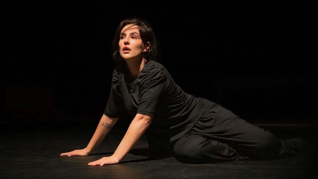 Kéfera de roupa preta sentada em chão de palco de teatro, com expressão de espanto