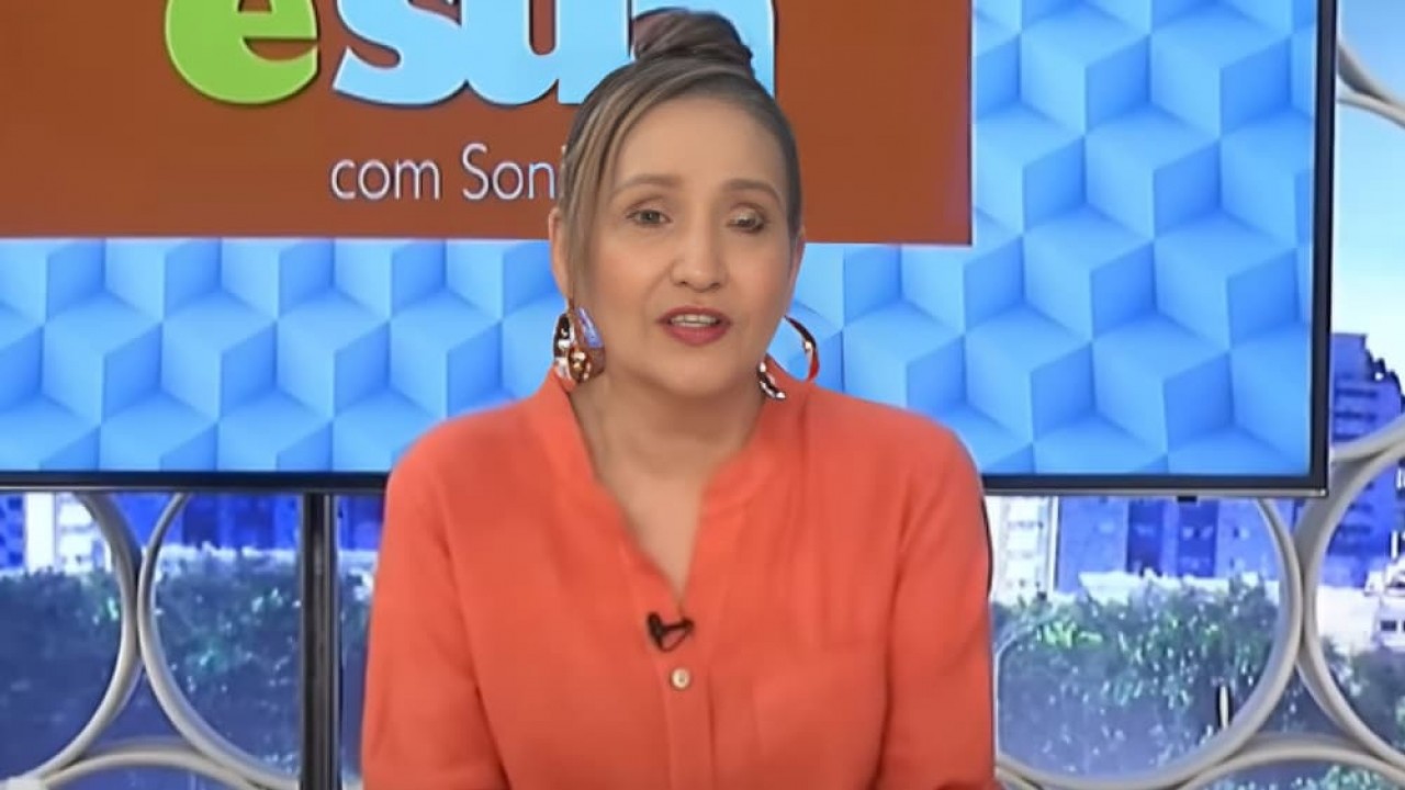 Sonia Abrão em edição do A Tarde é Sua, de roupa coral, falando, com cabelo preso em coque