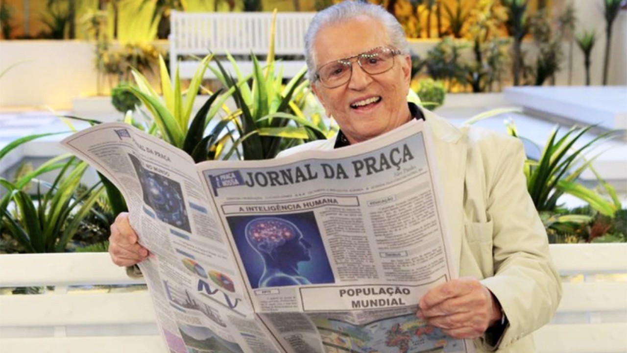 Carlos Alberto de Nóbrega no banco de A Praça É Nossa, segurando um jornal, sorridente