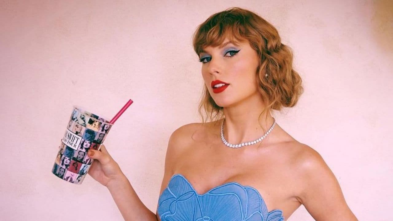 Taylor Swift de roupa tomara-que-caia azul, posando sem sorrir e segurando copo da The Eras Tour