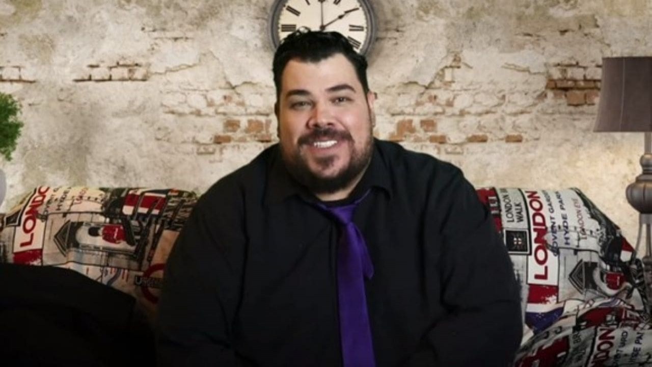 Daniel Araújo de camisa preta e gravata azul, sorrindo para a câmera em cenário colorido
