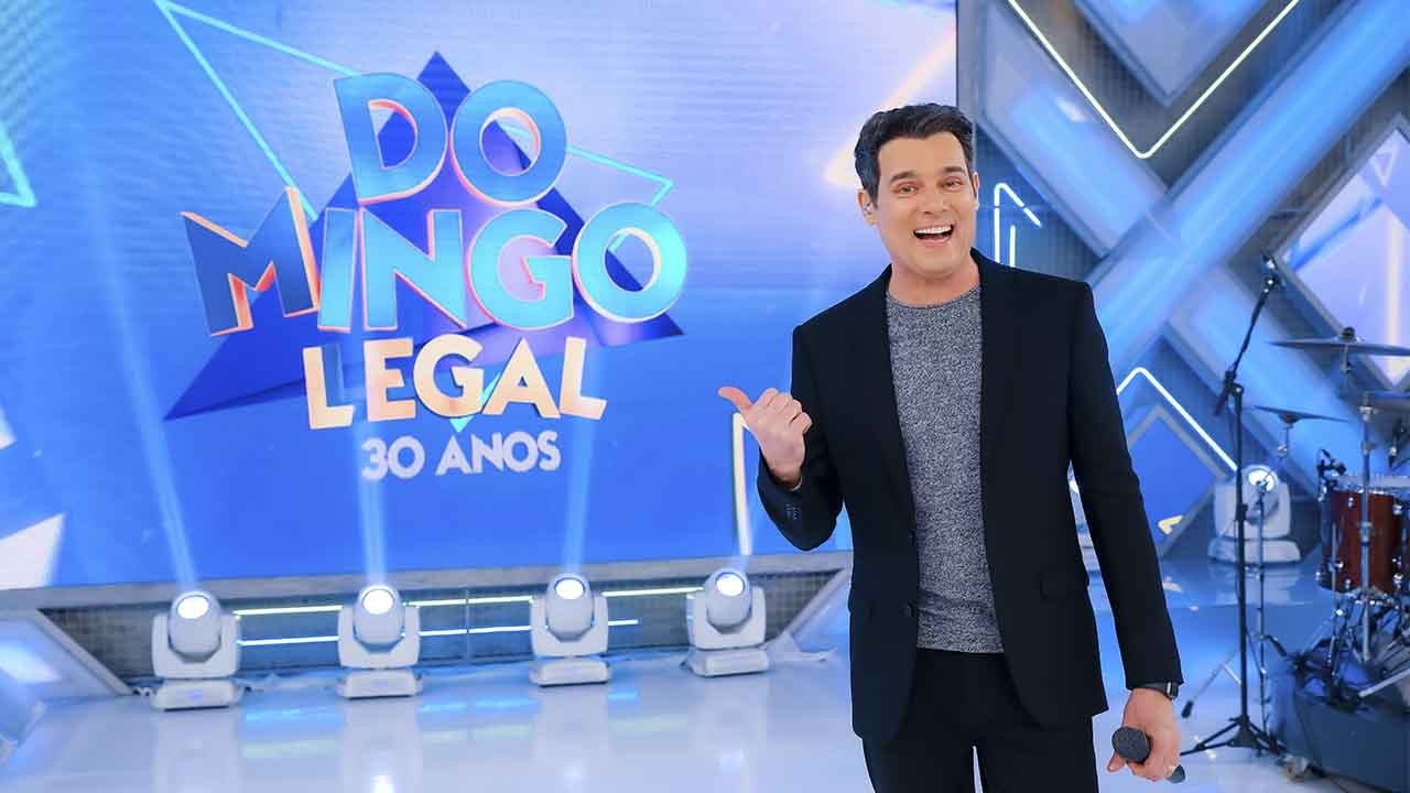 Celso Portiolli no palco do Domingo Legal posando na frente do telão com o logotipo do programa