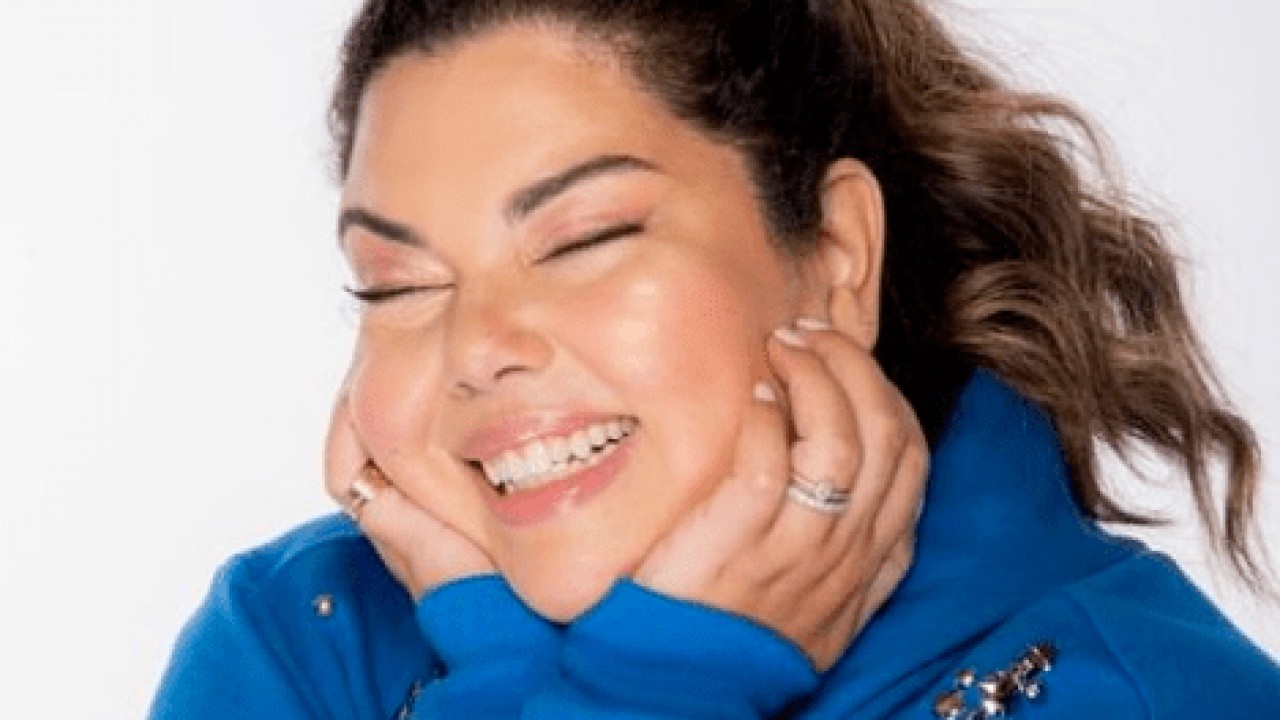 Fabiana Karla com roupa azul, mãos no rosto e sorrindo