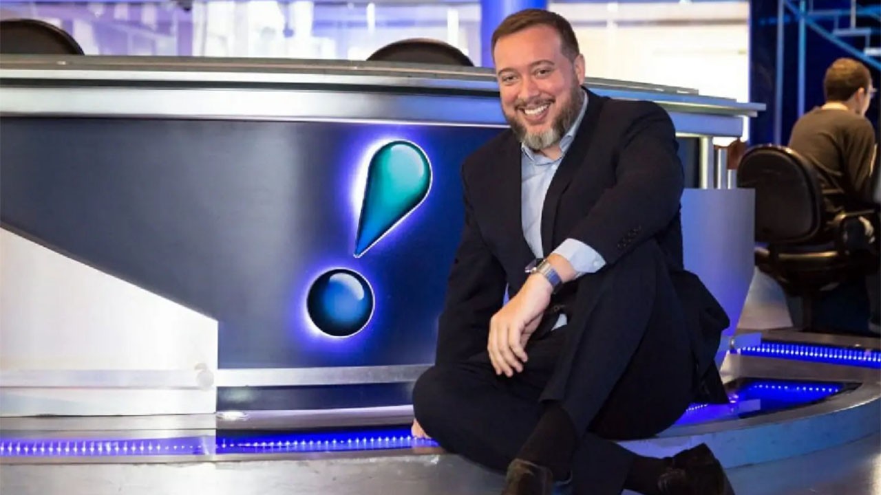 Franz Vacek sentado na frente de uma bancada com o logo da RedeTV!