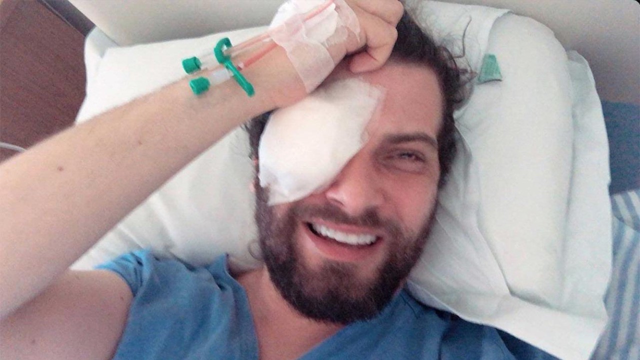 Saulo Meneghetti sorrindo em leito hospitalar com tampão no olho e mão na cabeça