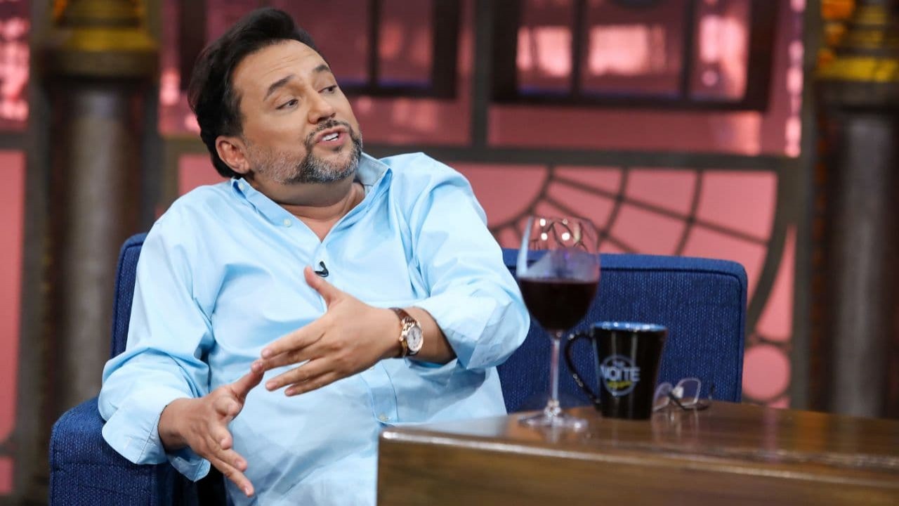 Geraldo Luís de camisa social azul, sentado falando e gesticulando no cenário do The Noite
