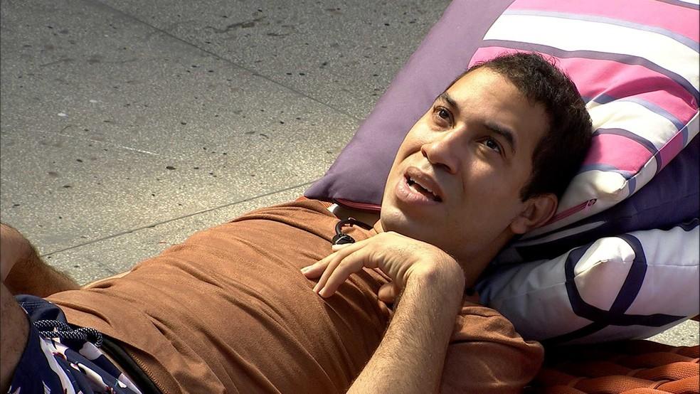 Gilberto está deitado em almofadas na área externa do BBB21 com a mão no peito e conversando