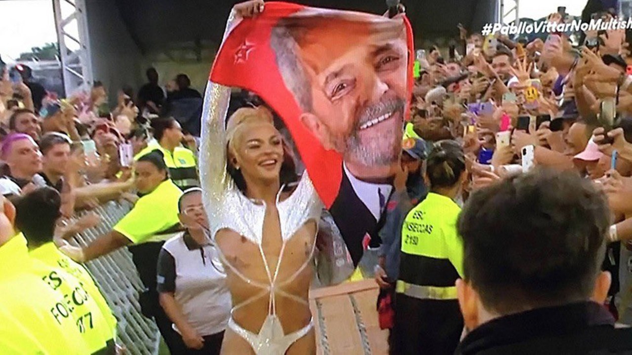 Pabllo Vittar próximo ao público, segurando toalha em que aparece estampado o rosto de Lula