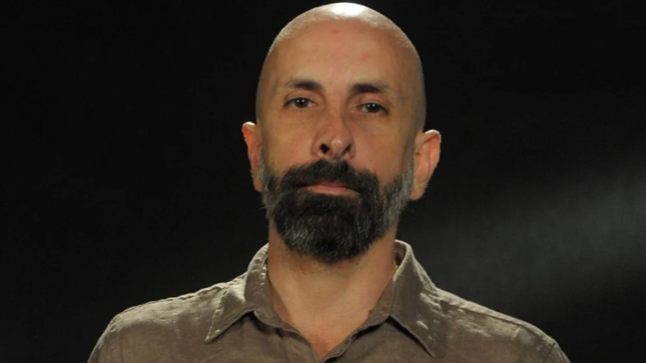 O autor João Ximenes Braga sério usando blusa marrom e à frente de fundo preto
