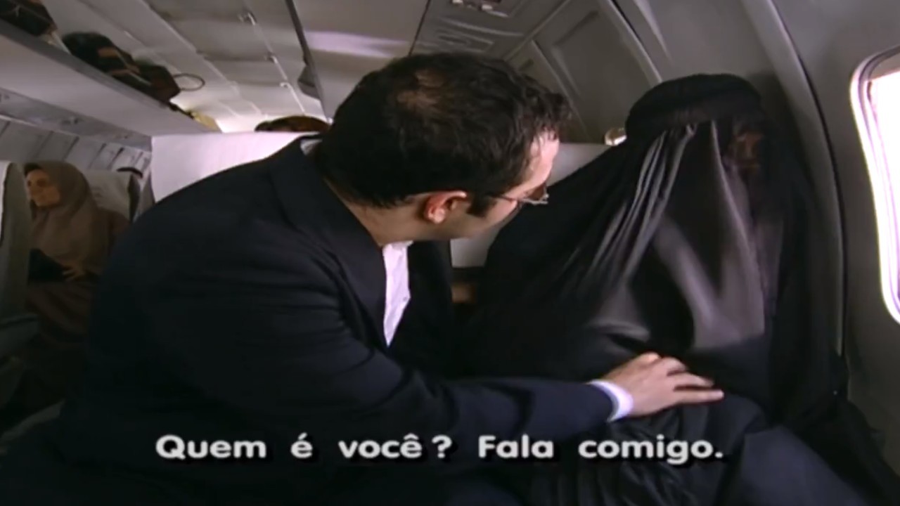 Jade sentada com véu preto ao lado de homem no avião em cena de O Clone