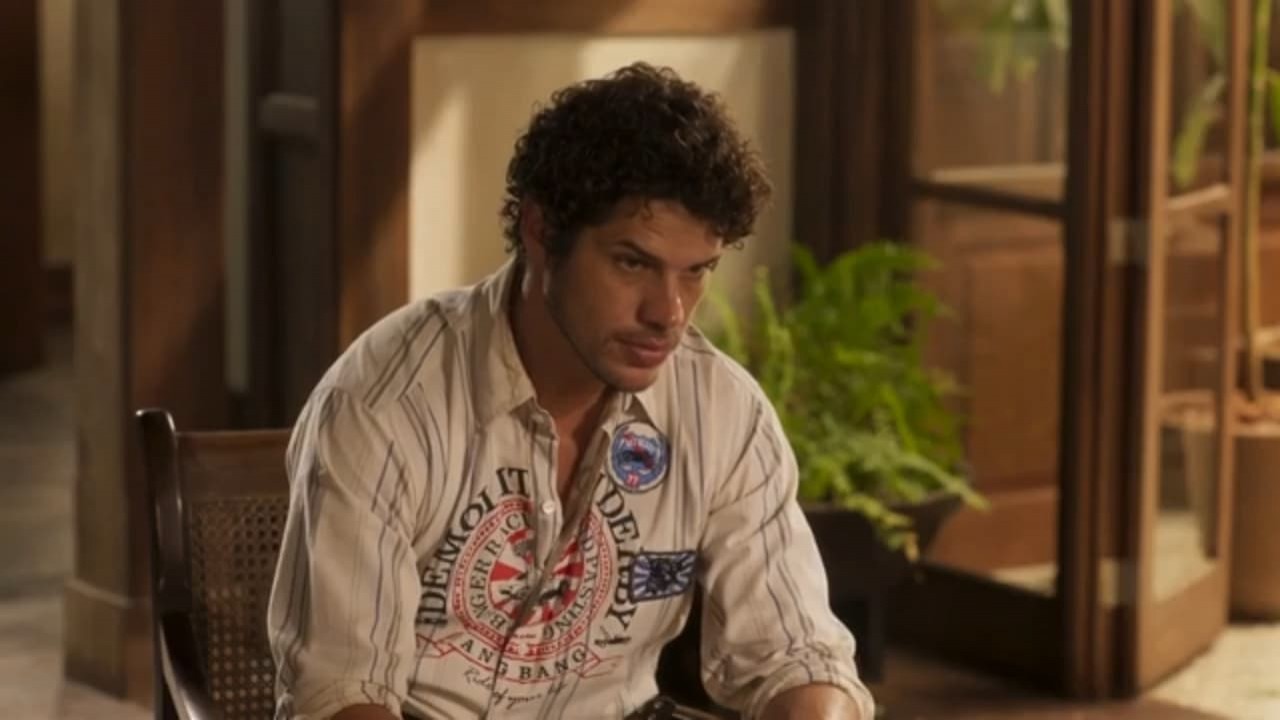 José Loreto como Tadeu em Pantanal. Ele está sentado na sala da fazenda